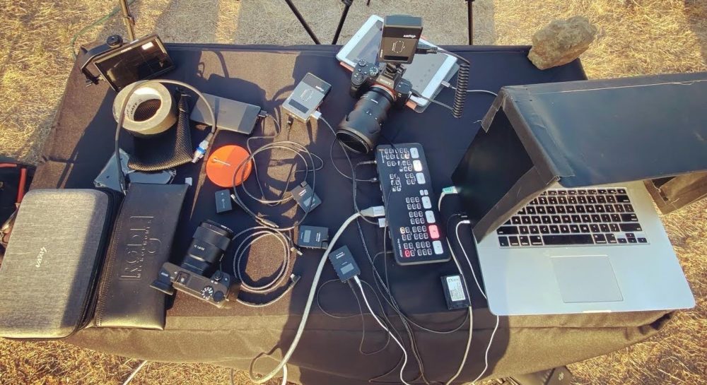 Live streaming equipment on a table. Video and audio gear and a laptop. Lots of cables and various equipment.