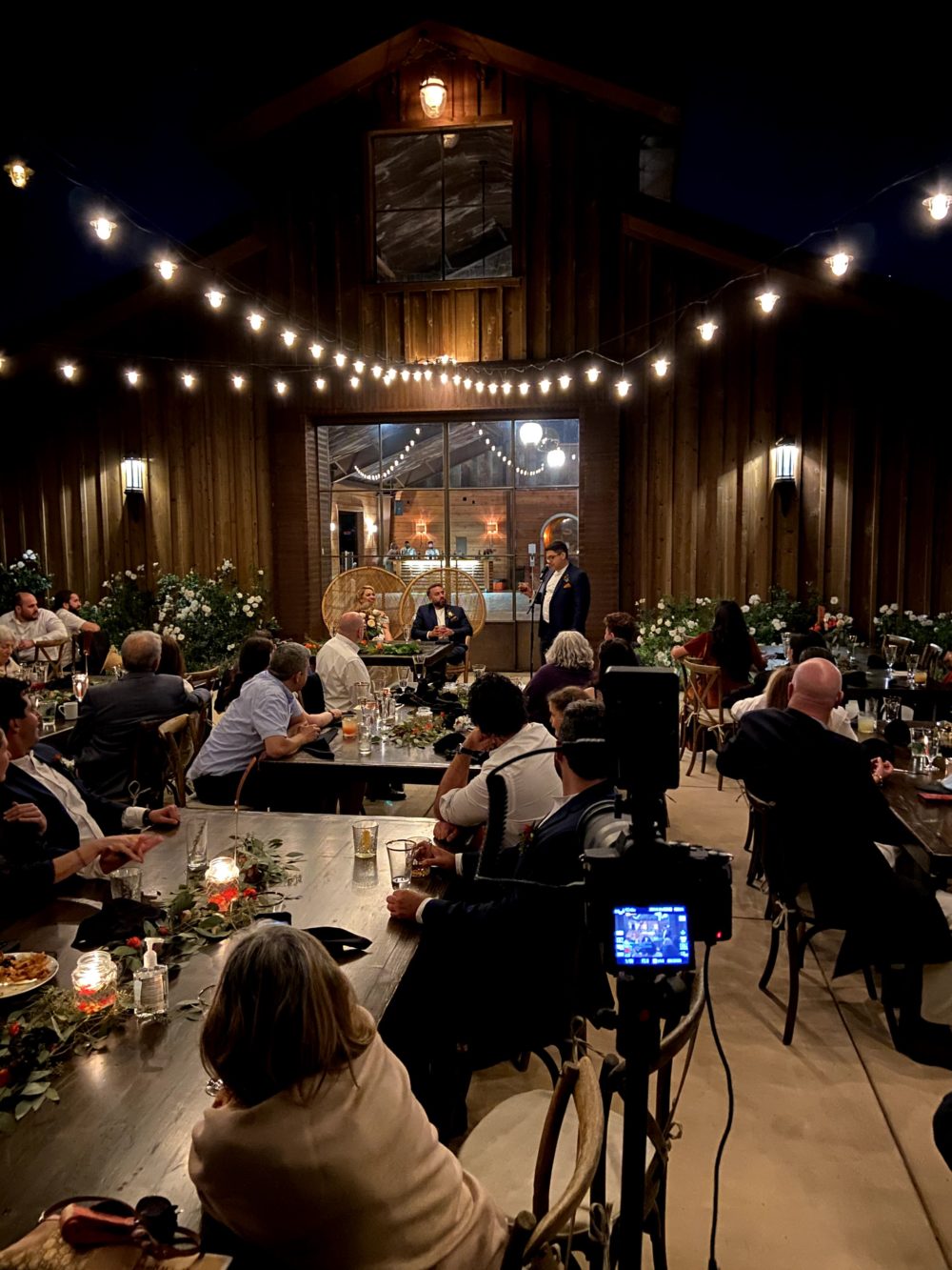 Evening outdoors at dining tables as guests watch best man giving toast to bride and groom sitting at main table in front of modern barn. 